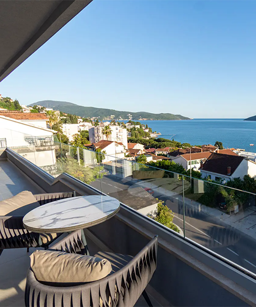 Herceg Novi'de Adriyatik Denizi'ne bakan bir balkonun cam parmaklıkları ardında, mermer desenli yuvarlak masa ve minderli iki sandalyenin bulunduğu huzurlu bir Karadağ körfez manzarası belirgin; sahilde sıralı beyaz evler, yeşil tepeler ve masmavi gökyüzüyle çevrili berrak denizin tadını çıkarıyor.