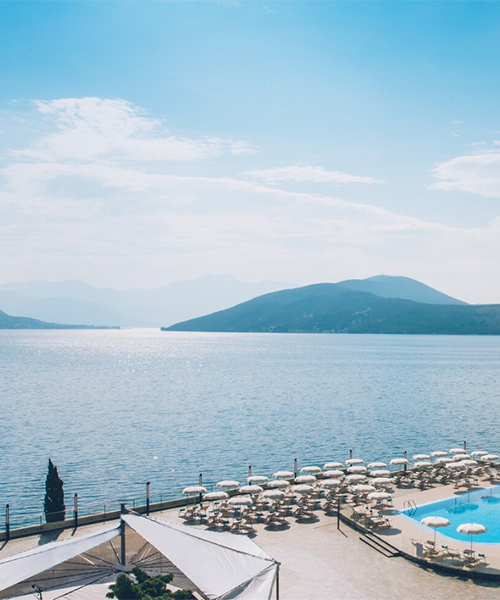 Güneşli bir Karadağ gününde, berrak Adriyatik Denizi'nin kıyısında yer alan lüks bir tatil köyünün geniş özel plaj alanı; uzakta dağ silüetleri, beyaz şemsiyelerle donatılmış şezlonglar, ışıldayan mavi bir yüzme havuzu ve büyük beyaz bir gölgelik yapısı ile huzurlu bir seyahat deneyimi sunuyor.