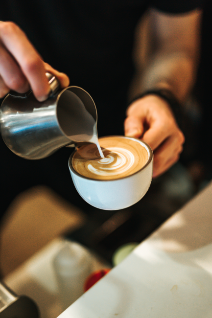 Bir barista, beyaz bir fincandaki kahvenin üzerine metal sürahiden köpüklü sütü özenle dökerek göz alıcı bir latte art deseni oluşturuyor ve böylece kahve nasıl yapılır sorusunun zarif bir yanıtını görsel olarak sunuyor.