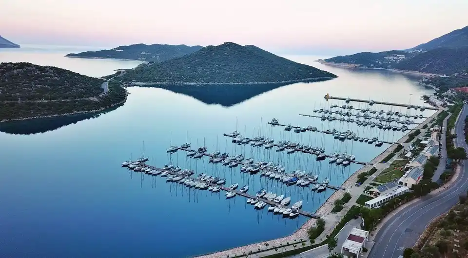 Alacakaranlıkta havadan çekilen bu fotoğraf, Akdeniz'in dingin sularında demirlemiş çok sayıda yelkenli ve motoryatla dolu, çevresi yemyeşil dağlar ve sahil şeridindeki modern tesislerle çevrili, Türkiye'nin en güzel marinalarından biri olan Kaş Marina'yı kuşbakışı gösteriyor.