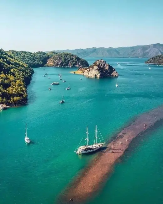Kızılkum Koyu'nda berrak turkuaz sularla çevrili, yeşil ağaçlarla kaplı tepelerin arasında demirlemiş çok sayıda tekne ve iki direkli büyük bir yelkenlinin uzanan sığ kırmızı kum patikasının yanında görüldüğü, güneşli bir havada çekilmiş nefes kesici kuşbakışı manzara.