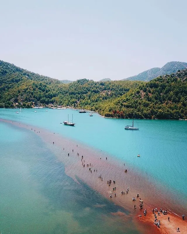 Marmaris seyahat rehberi kapsamında öne çıkan Kızkumu Plajı'nda, sahilden denize doğru uzanan kızıl kumlu patika üzerinde yürüyen ziyaretçiler, etrafı saran yemyeşil tepeler ve turkuaz sularda demirlemiş tekneler eşliğinde eşsiz bir Ege manzarası sunuyor.
