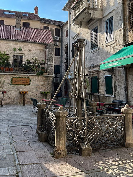 Güneşli bir Karadağ gününde, Kotor'un Arnavut kaldırımlı bir meydanında yükselen Karampa Kuyusu adı verilen tarihi su tulumbası, çevresindeki yeşil panjurlu eski taş binalar ve küçük kafelerle birlikte şehrin köklü geçmişini ve samimi Adriyatik atmosferini vurguluyor.