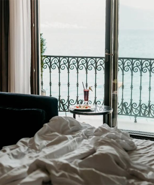 Konforlu bir yatağın ve şık bir içecek setinin bulunduğu otel odasından, Adriyatik'in berrak sularına nazır demir ferforje balkon korkulukları ardında huzurlu bir Karadağ deniz manzarası.