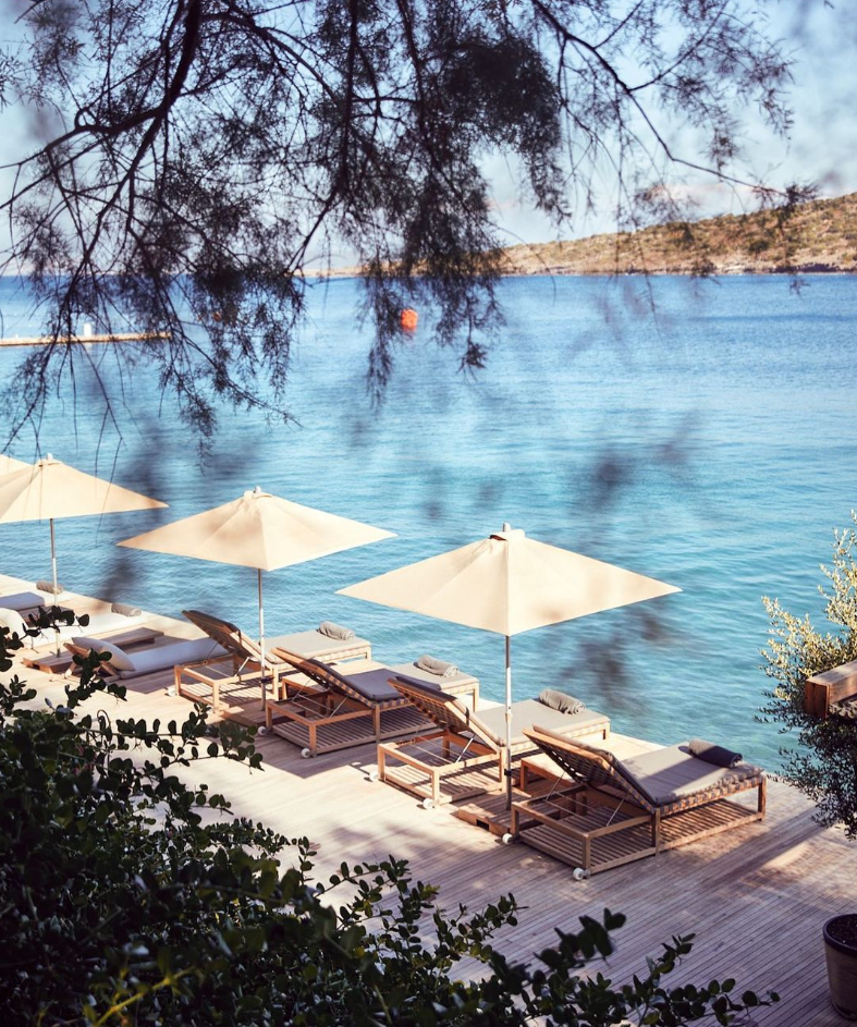 Bodrum'un göz kamaştıran Ege mavisinde, güneşin keyfini çıkarmak isteyenler için özenle düzenlenmiş, modern ahşap bir iskele üzerinde uzanan rahat şezlonglar ve onlara gölge sağlayan zarif bej şemsiyeler, yemyeşil dalların arasından sakin denize nazır huzurlu bir dinlenme alanı sunuyor.
