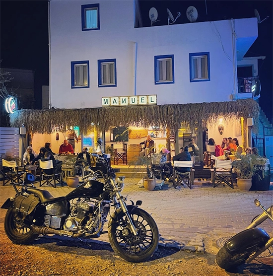 Bodrum'da gece vakti, önünde deri çantalı klasik bir motosikletin ve diğer motorların park ettiği, saz çatılı, ışıklı bir kafe veya barda dışarıda oturan müşterilerin olduğu hareketli bir ambiyans.