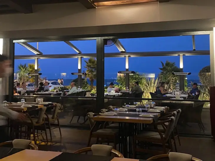 Samsun'un beğenilen restoranlarından birinin akşamüstü çekilmiş bu görüntüsünde, deniz kenarında konumlanan terasta palmiye ağaçları eşliğinde keyifli bir akşam yemeği yiyen misafirler ve iç mekandaki zarifçe düzenlenmiş masalar huzurlu bir atmosfer sunuyor.