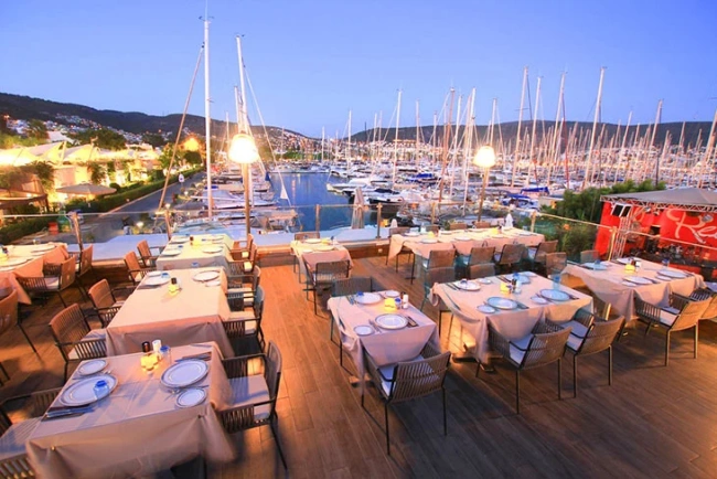 Alacakaranlıkta, Bodrum marinasının kalbindeki Marina Yacht Club'ın birçok yelkenli ve yatla dolu eşsiz manzarasına nazır, krem rengi masa örtüleri ve konforlu hasır sandalyelerle zarifçe düzenlenmiş terası, Bodrum'un en iyi restoranları arasında unutulmaz bir akşam yemeği deneyimi vadediyor.