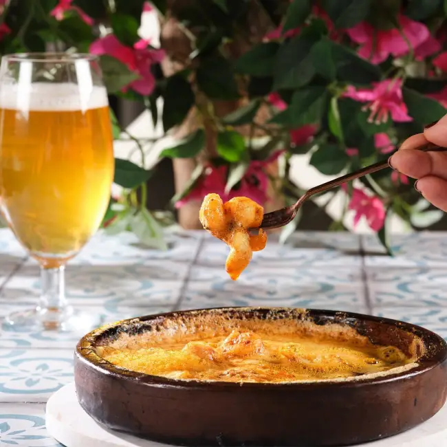 Samsun'un en beğenilen restoranlarından Müdavim'de, peynirli karides güveçten bir parça çatalla alınırken, arka planda serinletici bira bardağı ve canlı pembe çiçeklerle dolu hoş bir atmosfer göze çarpıyor.