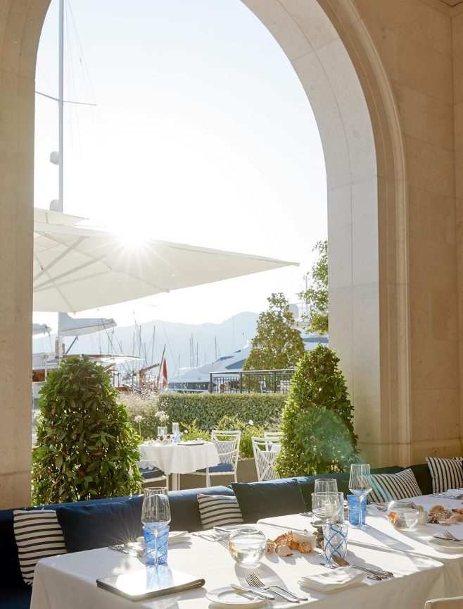 Taş kemerli bir restoranın dış oturma alanında, beyaz örtülü şık masalar ve mavi minderli banklar güneşte parlıyor; masalarda deniz kabukları ve özel cam eşyalar dikkat çekerken, arka planda Karadağ'ın lüks marinası Porto Montenegro'daki yelkenliler ve yatlar ile Adriyatik'in dağlık kıyı şeridi uzanıyor.