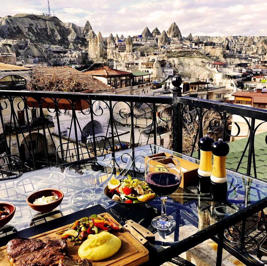 Kapadokya'nın eşsiz peri bacaları ve mağara evleriyle çevrili muhteşem manzarasına sahip bir balkonda, ızgara biftek, püre, sebze, salata ve bir kadeh kırmızı şarabın sunulduğu cam bir masa, bölgenin en iyi lezzet duraklarından birindeki keyifli bir yemeği betimliyor.