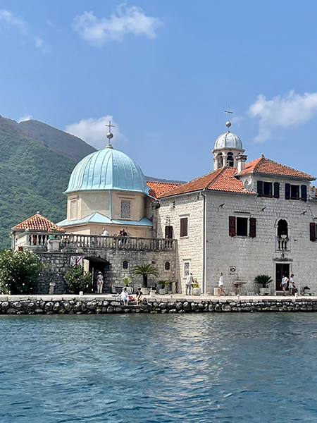 Perast açıklarında, masmavi Adriyatik sularının ortasında yükselen, tarihi taş duvarlara ve belirgin hafif mavi bir kubbesi olan kiliseye sahip, Karadağ'ın öne çıkan turistik noktalarından Our Lady of the Rocks Kayalar Üzerindeki Meryem Ana adası ve etrafında gezinen ziyaretçiler ile yemyeşil dağlarla çevrili berrak gökyüzü.