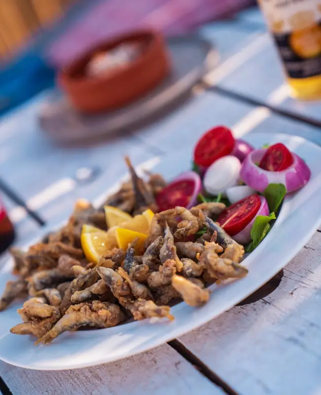 Ayvalık'ın nefis yemeklerinden biri olan, zeytinyağında kızarmış meşhur papalina tavası, yanında taze limon dilimleri, kırmızı soğan ve domateslerle süslenmiş beyaz bir tabakta, Kuzey Ege sofrasının davetkar atmosferini yansıtan ahşap bir masada iştah açıcı bir şekilde duruyor.
