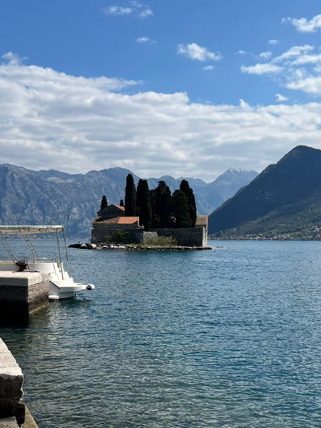 Karadağ'ın Adriyatik kıyılarındaki Perast'ta, Kotor Körfezi'nin berrak sularında yer alan, üzerinde tarihi bir kilise ve sık selvi ağaçlarının yükseldiği Sveti Đorđe Adası, sol önde iskeleye bağlı bir tekne ve ardındaki görkemli dağ manzaralarıyla bölgenin eşsiz doğal güzelliklerini sergiliyor.