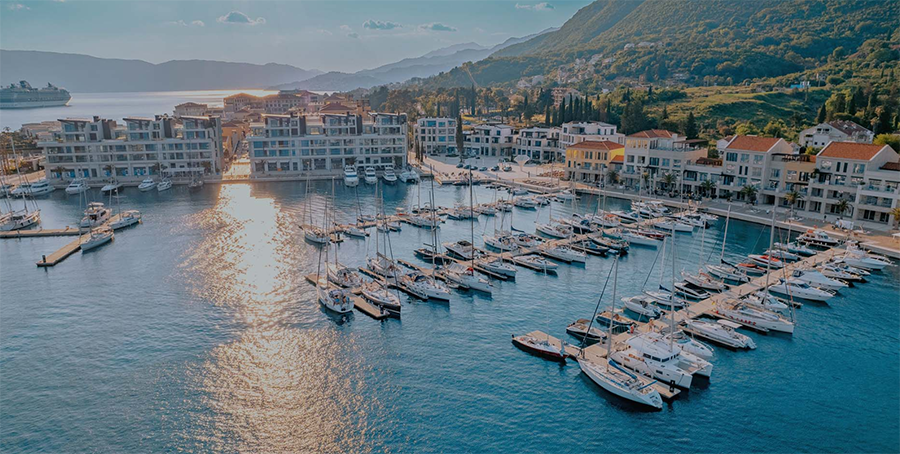 Güneşin parladığı bir Karadağ gününde, Adriyatik kıyısındaki Portonovi marinası, modern mimarili yapıların önünde masmavi sularda demirlemiş sayısız lüks yat ve tekneyle, yeşil dağların görkemli fonunda uzanıyor.