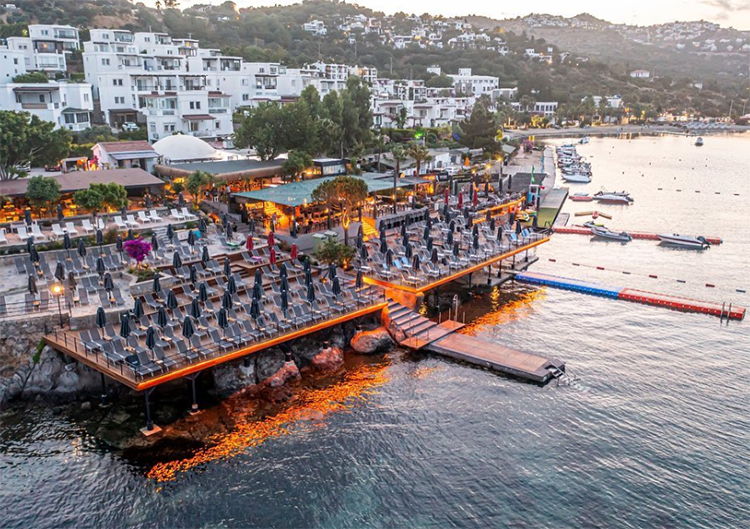 Bodrum'un Bitez'deki Sarnıç Beach'ini gösteren bu havadan görünümde, gün batımının veya şafağın yumuşak ışıkları altında, denizin üzerinde uzanan ahşap platformlardaki şezlonglar ve şemsiyeler, yamaçtaki beyaz evlerin eşlik ettiği Ege'nin mavisinde lüks bir sahil kulübü atmosferi sunuyor.