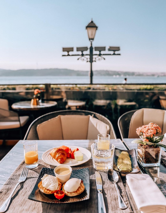 İstanbul'un göz alıcı Boğaz manzarasına sahip şık bir teraslı mekanda, güneşli bir sabah kahvaltısı; masada özenle hazırlanmış eggs benedict, kruvasan, taze meyveler, portakal suyu ve su, huzurlu bir başlangıcı işaret ediyor.
