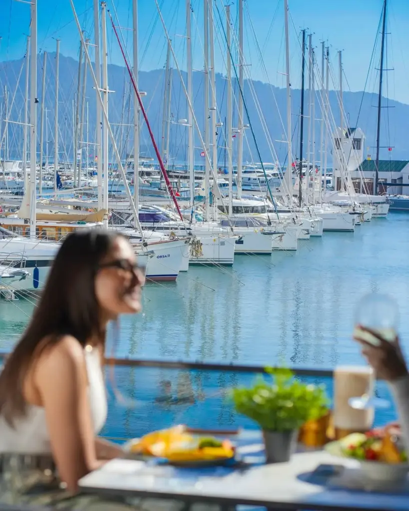 Güneşli bir günde, birçok yelkenli teknenin yan yana demirlediği hareketli bir marinada, ön planda bulanık görünen iki kişi yemek yiyerek Türkiye'nin güzelliğini ve marinaların sunduğu keyifli yaşamı deneyimliyor.