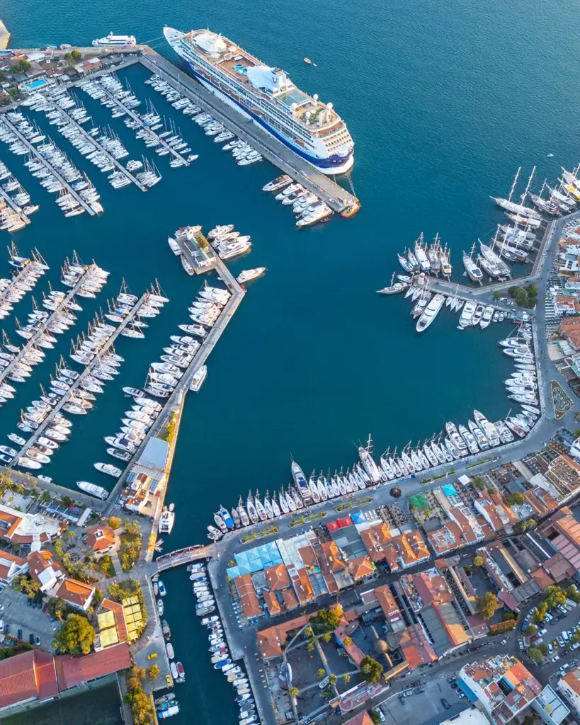 Türkiye'nin Ege ve Akdeniz kıyılarındaki hareketli bir marinayı kuşbakışı gösteren bu geniş fotoğrafta, uzun iskelelere sıralanmış sayısız yat ve yelkenli teknenin yanı sıra, limanın bir ucunda devasa bir kruvaziyer gemisi dikkat çekiyor ve çevresindeki canlı kıyı kasabası, bu bölgenin sunduğu tatil imkanlarını vurguluyor.