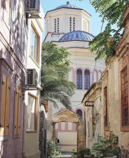 Dar, güneşli bir taş sokak, eski Rum evlerinin ve yeşil bitkilerin arasından, Ayvalık'ın tarihini yansıtan Cunda Adası'ndaki görkemli, krem rengi Taksiyarhis Kilisesi'ne ve belirgin kubbelerine doğru uzanıyor.