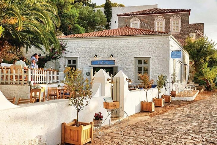 Hydra Adası'nda yer alan Téchnē restoranının beyaz badanalı, kırmızı kiremit çatılı binası, taş döşeli sokağa bakan ahşap masa sandalyeli açık hava terasıyla Yunan Adaları'nda tekneyle ulaşılabilen şirin bir deniz kıyısı mekânı olarak huzurlu bir atmosfer sunuyor.
