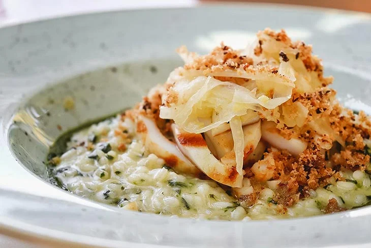 Yunan Adaları'nda tekneyle gidebileceğiniz enfes bir restoran olan Téchnē'den, yeşil otlarla zenginleştirilmiş kremalı risotto üzerinde ızgara kalamar dilimleri, ince rendelenmiş soğan benzeri sebzeler ve çıtır ekmek kırıntılarıyla sunulan, Akdeniz mutfağının etkileyici bir örneği olan deniz ürünlü özel bir lezzet.