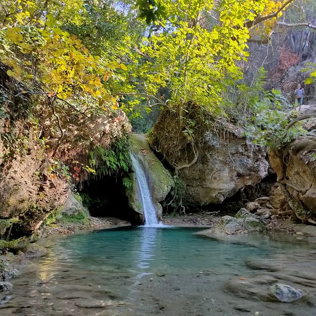 Marmaris seyahat rehberinin öne çıkan noktalarından Turgut Şelalesi, yemyeşil ağaçlar ve yosunlu kayalarla çevrili alanda berrak turkuaz rengi suyuyla birikinti havuzuna dökülürken, uzakta kayalık bir patikada duran bir ziyaretçi doğanın huzur veren bu dinginliğini izliyor.