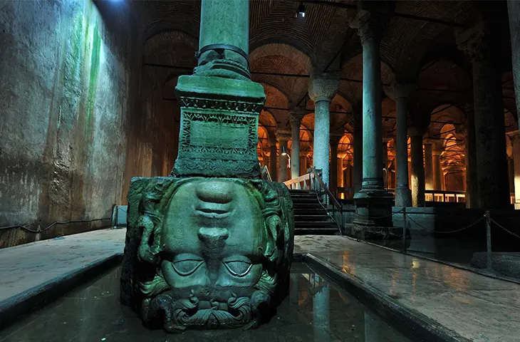 Yerebatan Sarnıcı'ndaki Medusa başının ters çevrilmiş haliyle gösterildiği geniş açılı bir fotoğraf. Baş, suyun içinde bir kaide üzerinde duruyor ve etrafındaki sütunlar, tavan ve mistik aydınlatma yapının büyüleyici atmosferini ortaya koyuyor.