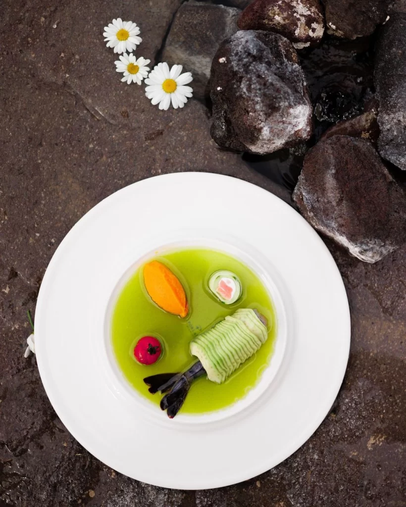 Kapadokya'da ziyaret edilebilecek gastronomik mekanların birinde servis edilen, açık yeşil bir sosun içinde özenle sunulan, ince dilimlenmiş salatalıkla sarılı büyük bir karidesin yanı sıra turuncu bir püre ve renkli garnitürlerle süslenmiş, arka planda ıslak koyu renkli taşlar ve papatyaların görüldüğü zarif bir tabak.