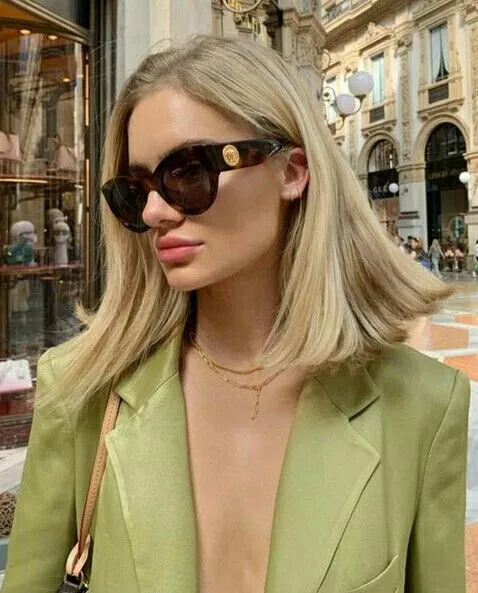 Şık kahverengi güneş gözlükleri ve zarif altın kolyeleriyle açık yeşil bir blazer giyen sarışın bir kadın, omuzlarına dökülen doğal dalgalı küt saç modeliyle tarihi bir çarşının vitrinleri önünde modern bir duruş sergiliyor.