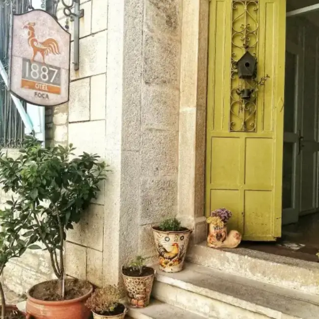 Foça Hotel 1887'nin taş bloklu duvarı önünde, üzerinde horoz figürü ve otel adının yazılı olduğu tabelasıyla, kuş evi detaylı parlak sarı kapısı ve basamaklarında horoz desenli saksılar içinde canlı bitkilerle süslü davetkar girişi, Ege'nin bu şirin Foça rehberi kapsamında dikkat çekiyor.