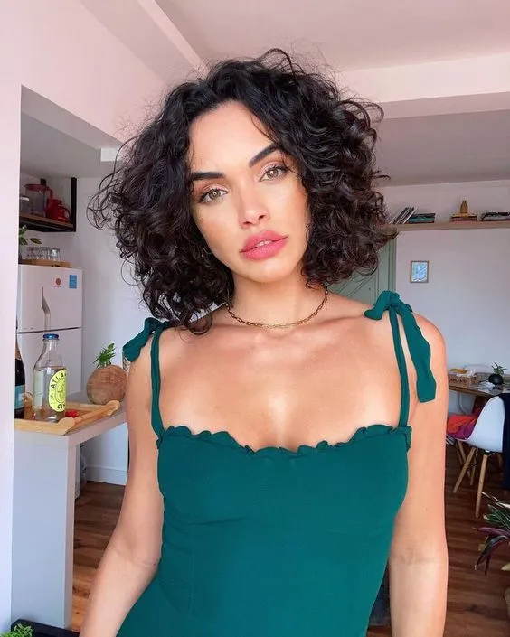 Dolgun ve omuz hizasında kıvırcık küt saç modeline sahip, canlı bakışları ve pembe rujuyla dikkat çeken, yeşil askılı bluz giymiş bir kadın, makyajıyla modern ve şık bir görünüm sergiliyor.