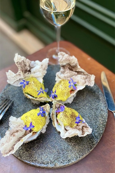 Fransız mutfağının ve Paris'in gastronomi zenginliğinin bir göstergesi olarak, Frenchie logosu görünen bir kadeh beyaz şarap eşliğinde, üzerinde parlak sarı sos, koyu renkli baharat serpiştirilmiş çıtır garnitürler ve narin mor çiçeklerle süslenmiş dört adet taze istiridye koyu gri bir servis tabağında şıkça sunuluyor.