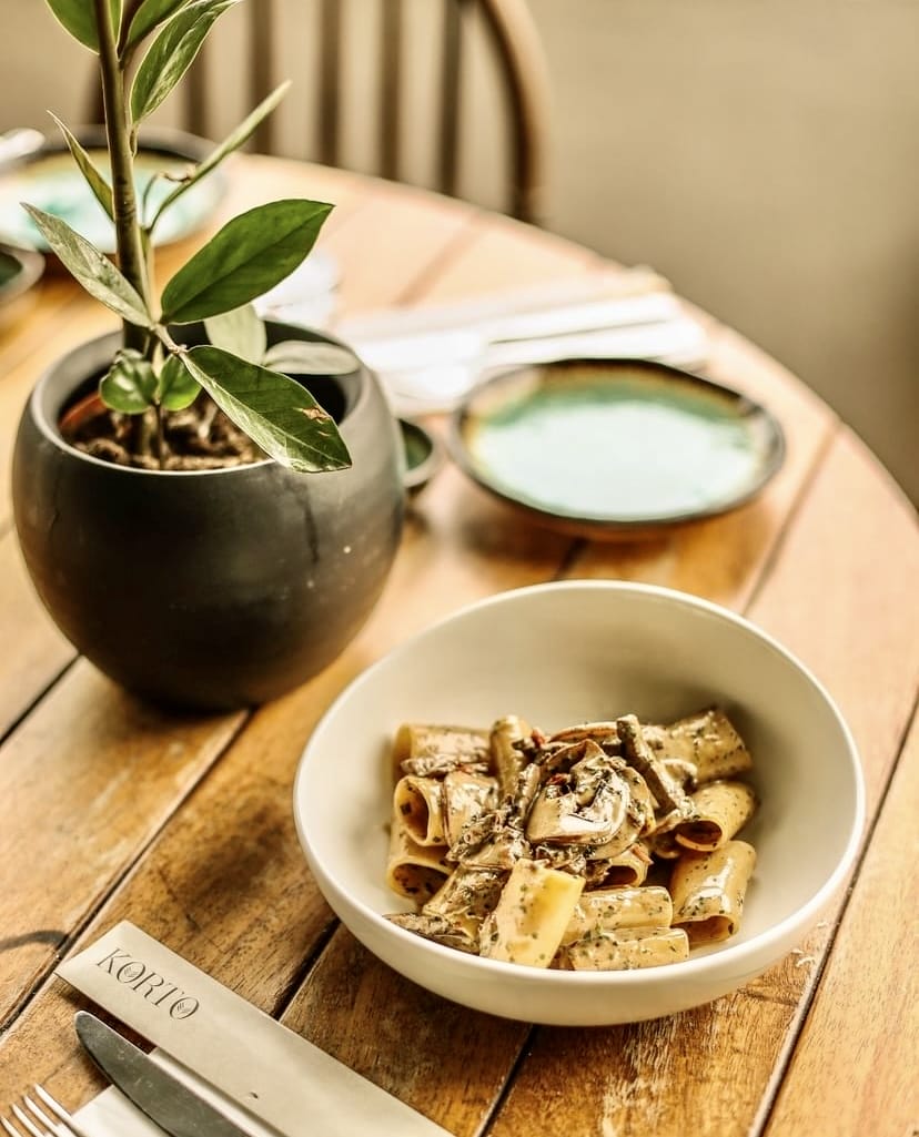 Beyaz bir tabakta, kremalı soslu ve mantarlı rigatoni makarna, ahşap bir masanın üzerinde, küçük yeşil bir bitki ve bulanık arka planda ek tabaklarla beraber sunulurken, İstanbul'un en iyi teraslı mekanlarından Korto'da lezzetli bir yemek deneyimini çağrıştırıyor.