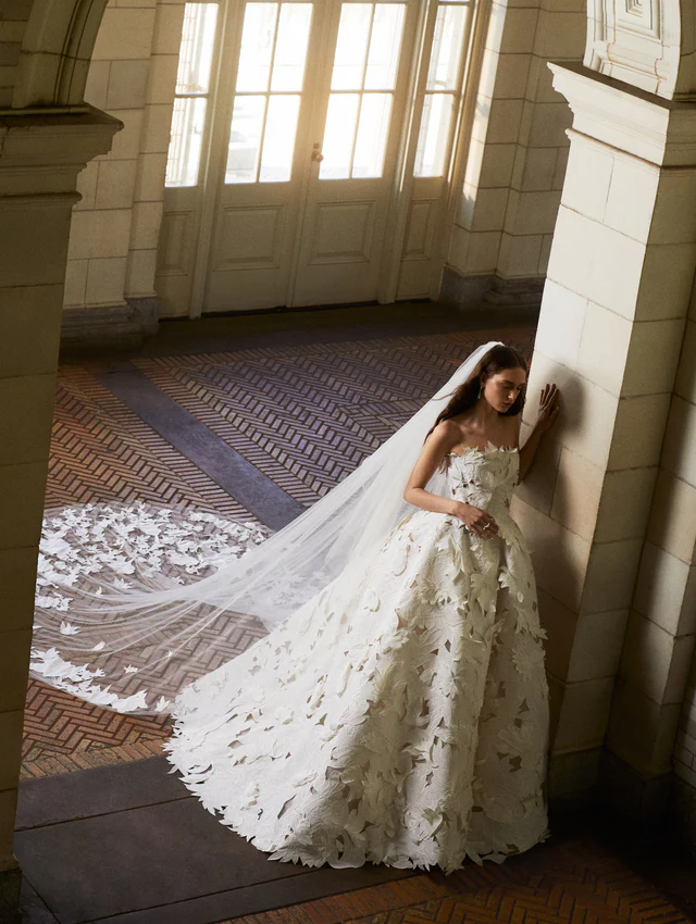 Oscar de la Renta 2025 koleksiyonundan, yaprak aplikeli straplez gelinlik ve uyumlu tül duvak detaylı model.