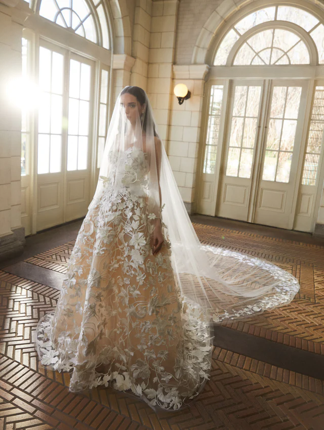 Oscar de la Renta 2025 koleksiyonundan, nude astarlı ve çiçek aplikeli tül gelinlik ile uzun dantel duvaklı model.