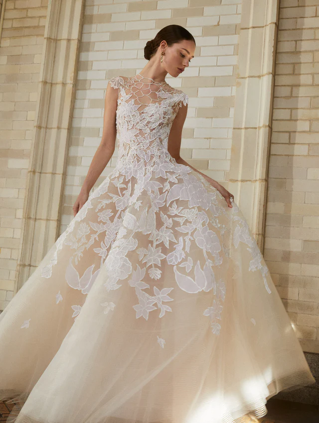 Oscar de la Renta 2025 koleksiyonundan, floral illüzyon yaka ve üç boyutlu çiçek aplikeleriyle süslenmiş transparan tül gelinlik modeli.