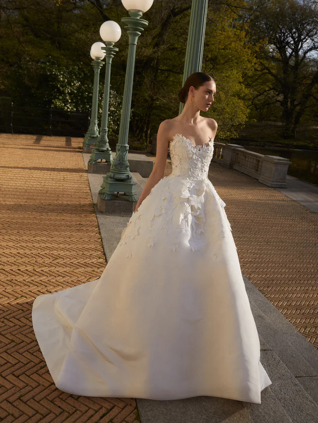 Oscar de la Renta 2025 koleksiyonundan, straplez yaka ve çiçek aplikeli klasik prenses kesim gelinlik modeli.