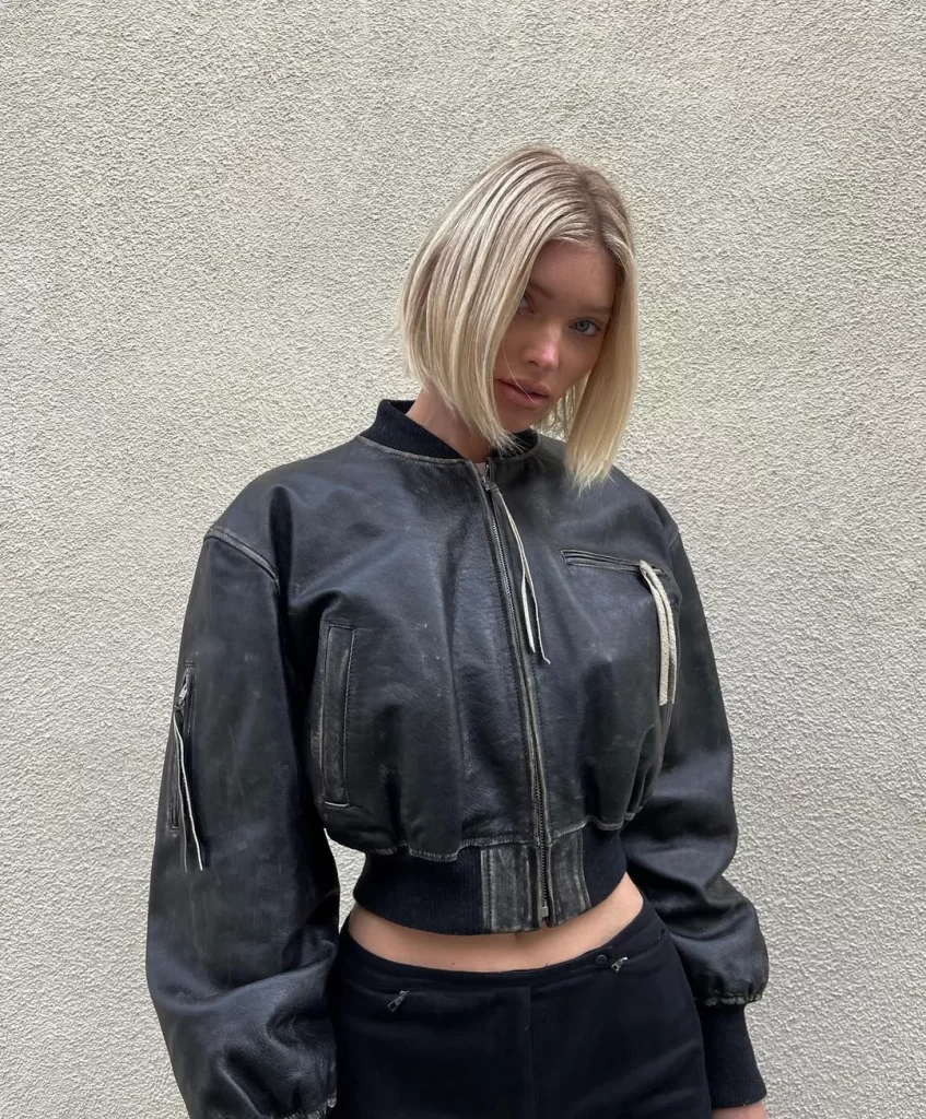 Elsa Hosk, pürüzsüz platin sarısı, çene hizasındaki modern kısa küt saç modeliyle, sade dokulu bir duvar önünde dururken şık siyah yıpranmış bomber ceketini giymiş, objektife kararlı bir ifadeyle bakıyor.