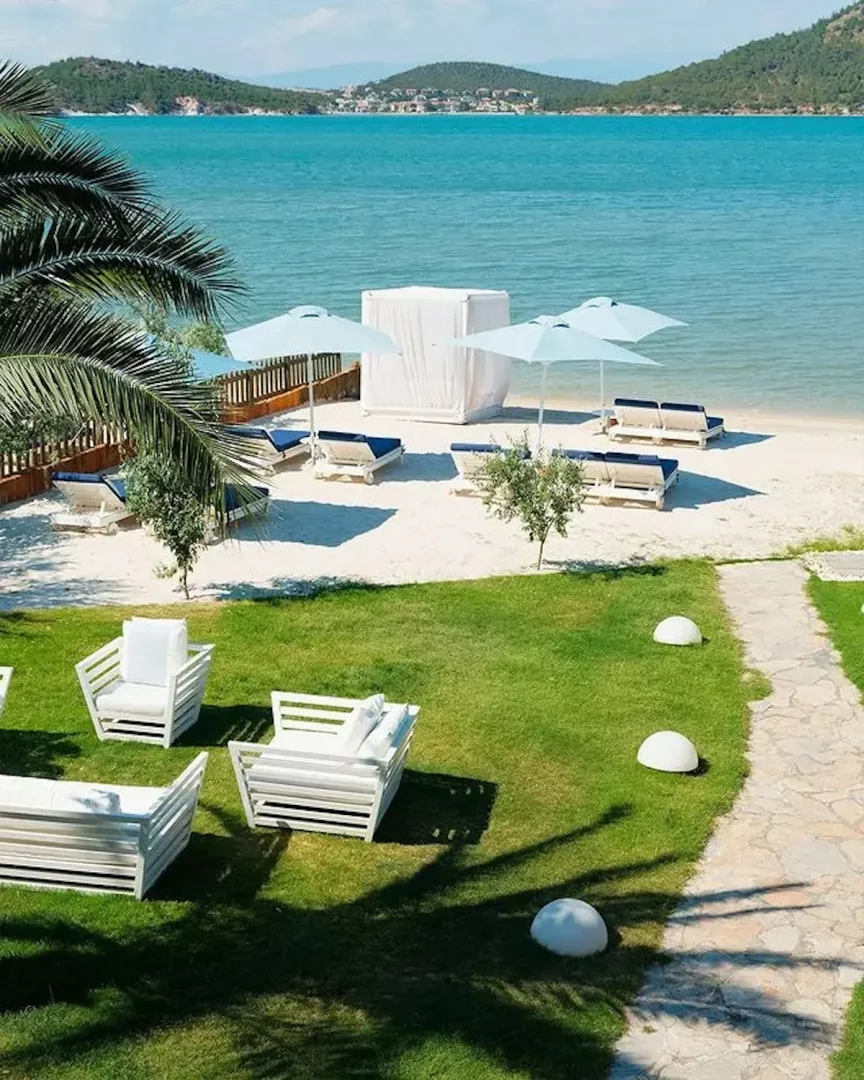 Ayvalık D Resort Murat Reis evcil hayvan dostu otelin güneşli kumsalında mavi şemsiyeler altında sıralanmış şezlonglar, yemyeşil çimler üzerindeki beyaz modern oturma grupları ve masmavi deniz ile çevrili sakin bir tatil ortamı.