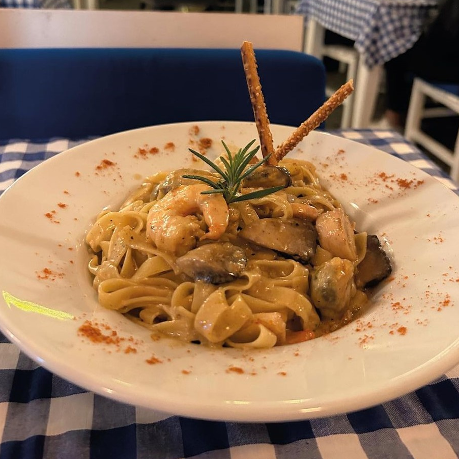 Mavi beyaz kareli bir masa örtüsü üzerinde sunulan, kremsi soslu karidesli ve mantarlı fettuccine tabağı, üzerindeki taze biberiye dalı ve iki çıtır tuzlu çubuk krakerle süslenmiş, tabağın kenarlarında ise kırmızı toz baharat serpintileriyle tamamlanmış bir ana yemek.