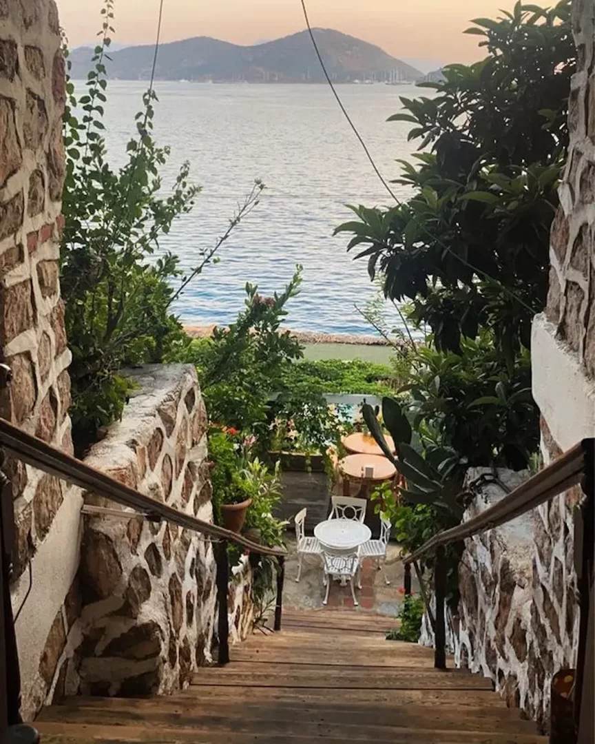 Rustik ahşap merdivenlerden aşağıya, yeşilliklerle çevrili taş duvarların arasından Bozburun Dolphin Hotel'in deniz manzaralı, beyaz masa ve sandalyelerin bulunduğu sakin bahçesine bakış; uzakta dağlar ve tekneler görülüyor.