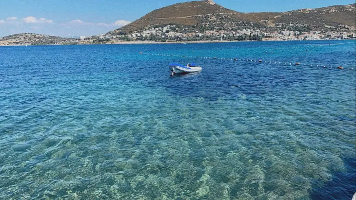 Masalımsı Foça'nın tertemiz ve berrak turkuaz rengi Ege sularında yüzen küçük bir botun gözüktüğü bu plaj görüntüsünde, şehrin dağlık kıyı şeridi ve yeşilliklerle kaplı tepeleri huzurlu bir tatil vaat ediyor.