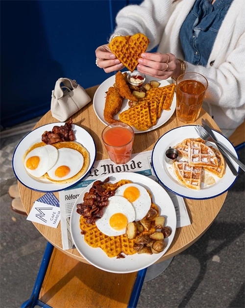 Cafe Mareva'nın dış mekanında, üzerinde kızarmış tavuk, waffle, patates, pastırma ve iki farklı yumurta tabağı bulunan yuvarlak bir masada, bir kişinin elinde kalpli bir waffle tuttuğu bu fotoğraf, menüsündeki glutensiz ve laktozsuz seçenekleriyle öne çıkan, En İyi Paris Restoranları listesindeki keyifli kafelerden birinde zengin bir brunch deneyimini gözler önüne seriyor.