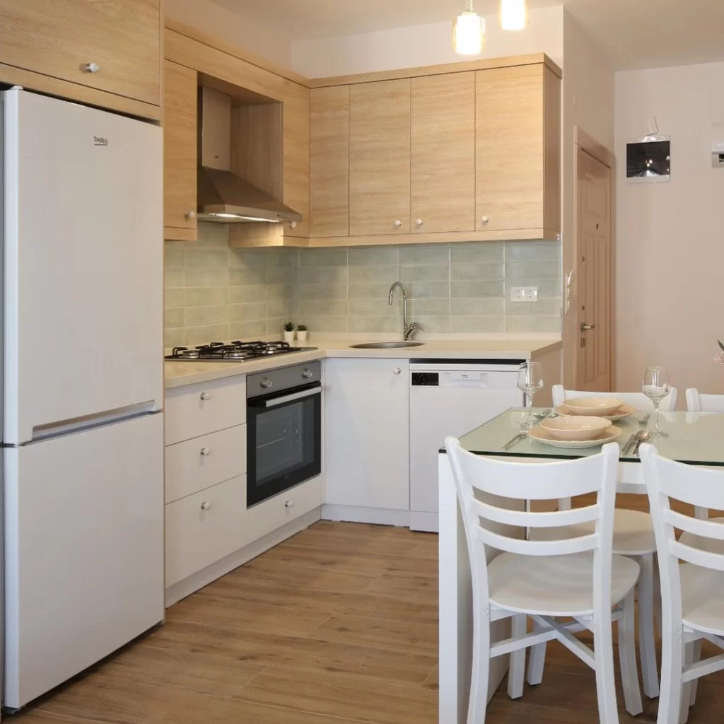 Modern ve aydınlık mutfak, açık renk ahşap üst dolaplar, beyaz alt dolaplar, beyaz Beko buzdolabı, ankastre fırın, ocak ve bulaşık makinesi gibi beyaz eşyalarla donatılmış olup, açık yeşil fayanslı tezgah arası ve ahşap görünümlü zeminiyle şık bir görünüm sunarken, sağda üzerinde tabaklar ve kadehler bulunan cam bir yemek masası ile beyaz sandalyeler dikkat çekiyor.