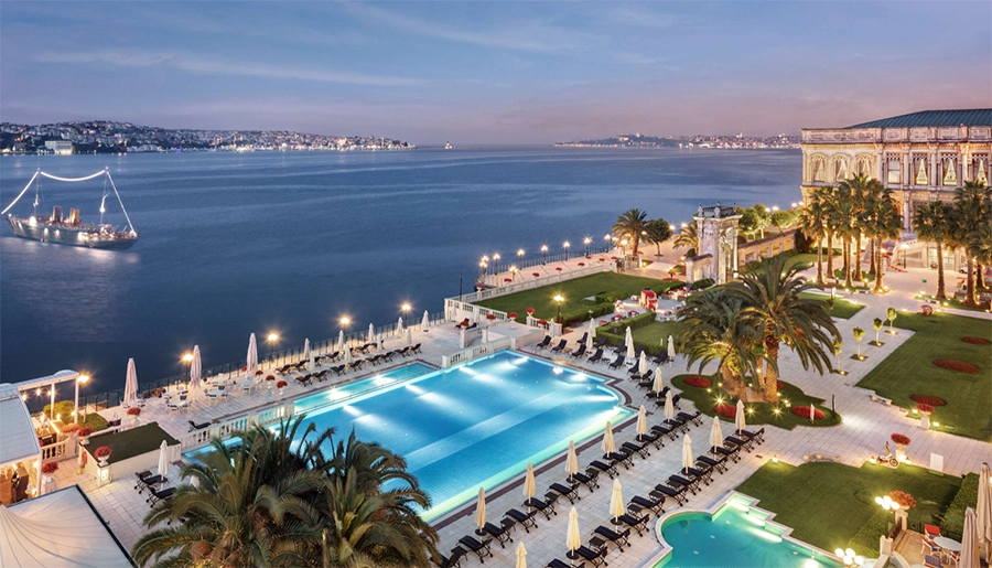Çırağan Sarayı Kempinski'nin Boğaz kenarında, akşam alacakaranlığında ışıklarla aydınlatılmış büyük açık yüzme havuzu, çevresindeki şezlonglar, palmiye ağaçları ve tarihi sarayın ışıltısıyla İstanbul'da lüks bir yaz kaçamağı atmosferi sunarken, denizde süzülen ışıklı bir gemi ve karşı kıyının silüeti eşsiz bir manzara oluşturuyor.