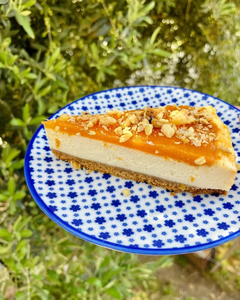 Mavi beyaz yıldız desenli seramik bir tabakta sunulan, kalın kremalı iç tabakası ve altında bisküvi tabanı bulunan, üzeri parlak turuncu meyve sosu ve bolca ceviz içiyle zenginleştirilmiş lezzetli bir cheesecake dilimi, bulanık yeşil bitki örtüsü arka planında iştah açıcı görünüyor.
