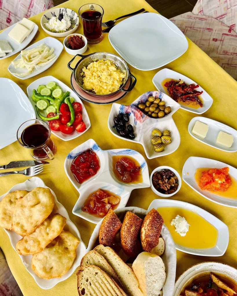 Sarı bir masa örtüsü üzerinde bol çeşitliliğe sahip, menemen, zeytin, peynirler, reçeller, tereyağı, kaymaklı bal, pişi, ekmek çeşitleri ve çay gibi birçok geleneksel yiyecek ve içeceği içeren zengin bir Türk kahvaltısı sofrası.