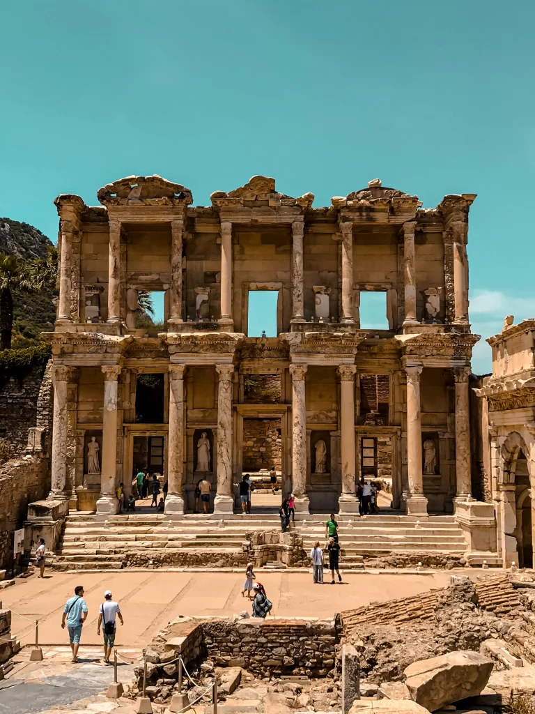 Efes Antik Kenti'nin kalbinde yer alan, görkemli iki katlı cephesi, detaylı sütunları ve nişlerindeki heykelleriyle büyüleyici bir yapı olan Celsus Kütüphanesi'nin önünde, merdivenlerde ve çevresinde dolaşan çok sayıda ziyaretçiyi gösteren, pırıl pırıl mavi bir gökyüzü altında çekilmiş geniş bir görüntü.