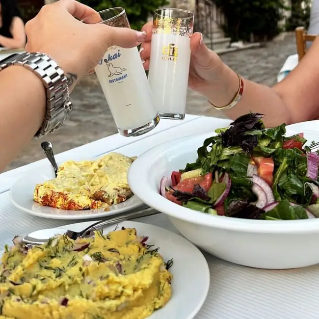 İki kişinin Foça Balık Restoranı'nın fok balığı logolu kadehi de dahil olmak üzere iki rakı kadehiyle kadeh kaldırdığı, Ege mutfağının zenginliğini yansıtan; taze yeşillik, domates, kırmızı soğanla hazırlanmış büyük bir salata kasesinin, dereotlu fava ve üzeri soslu başka bir mezenin beyaz masayı süslediği canlı bir an.